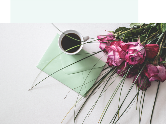 Some roses surround a green envelope and cup of coffee on a table